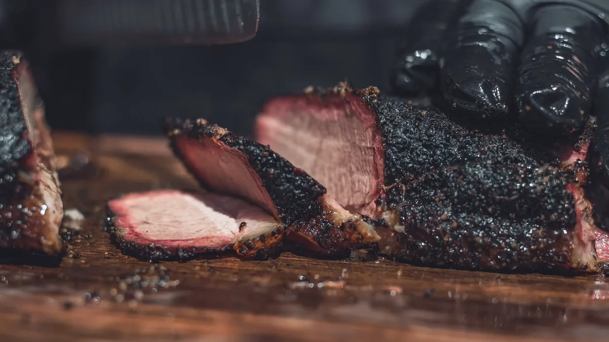 Gloved hands slicing a heavily peppered smoked brisket on a wooden board, showing the dark bark and pink smoke ring.