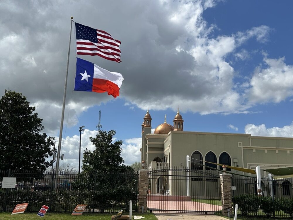 U.S. and Texas flags flying outside a mosque with political yard signs near the fence.