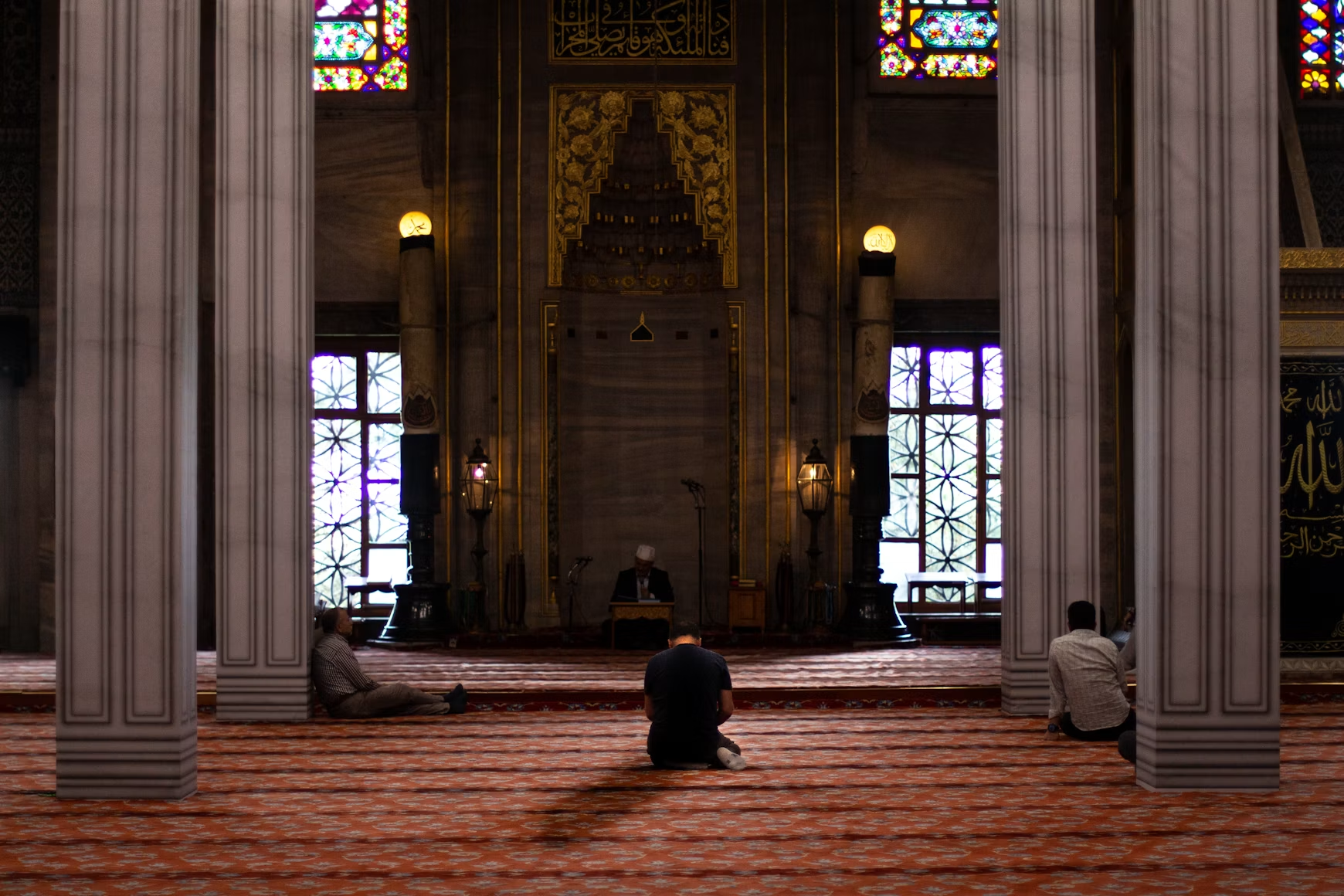Interior of a mosque with worshippers seated between tall columns and stained glass.