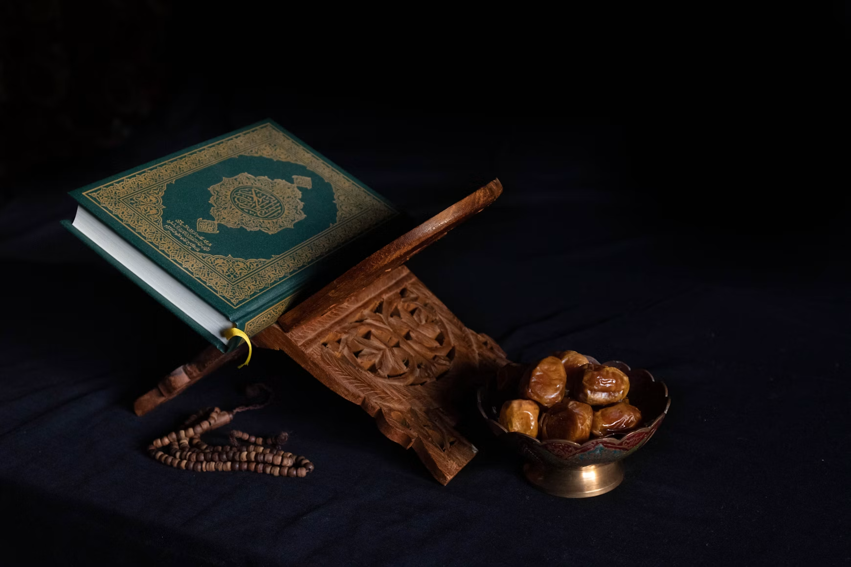 A Qur'an on a wooden stand beside dates and prayer beads on a dark cloth.