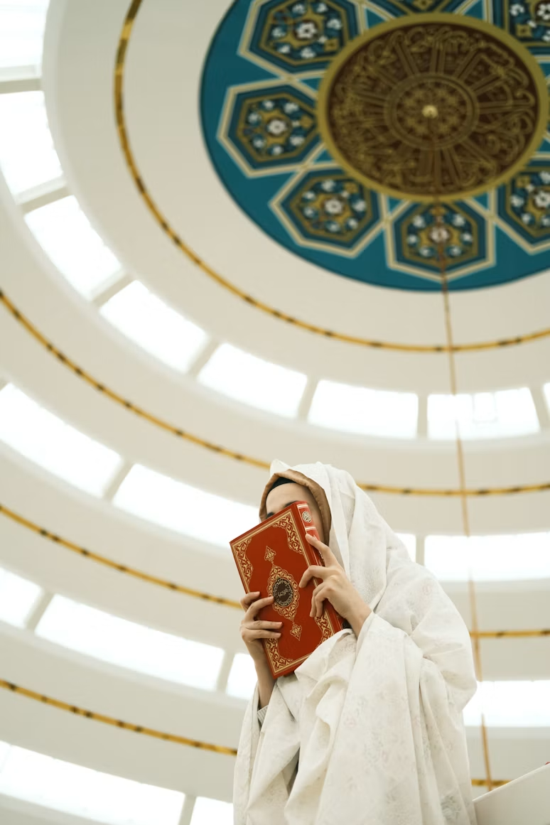 A person in white clothing holding a red Qur'an beneath a decorated mosque ceiling.