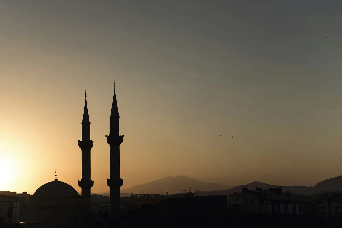 Two minarets silhouetted against an amber sky at sunset above a low skyline.