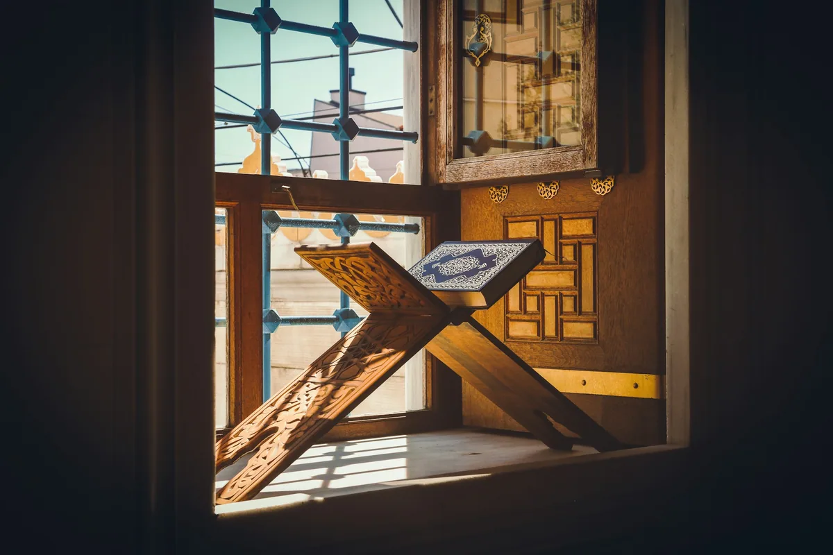A decorated Qur'an resting on a carved wooden stand beside a sunlit window.
