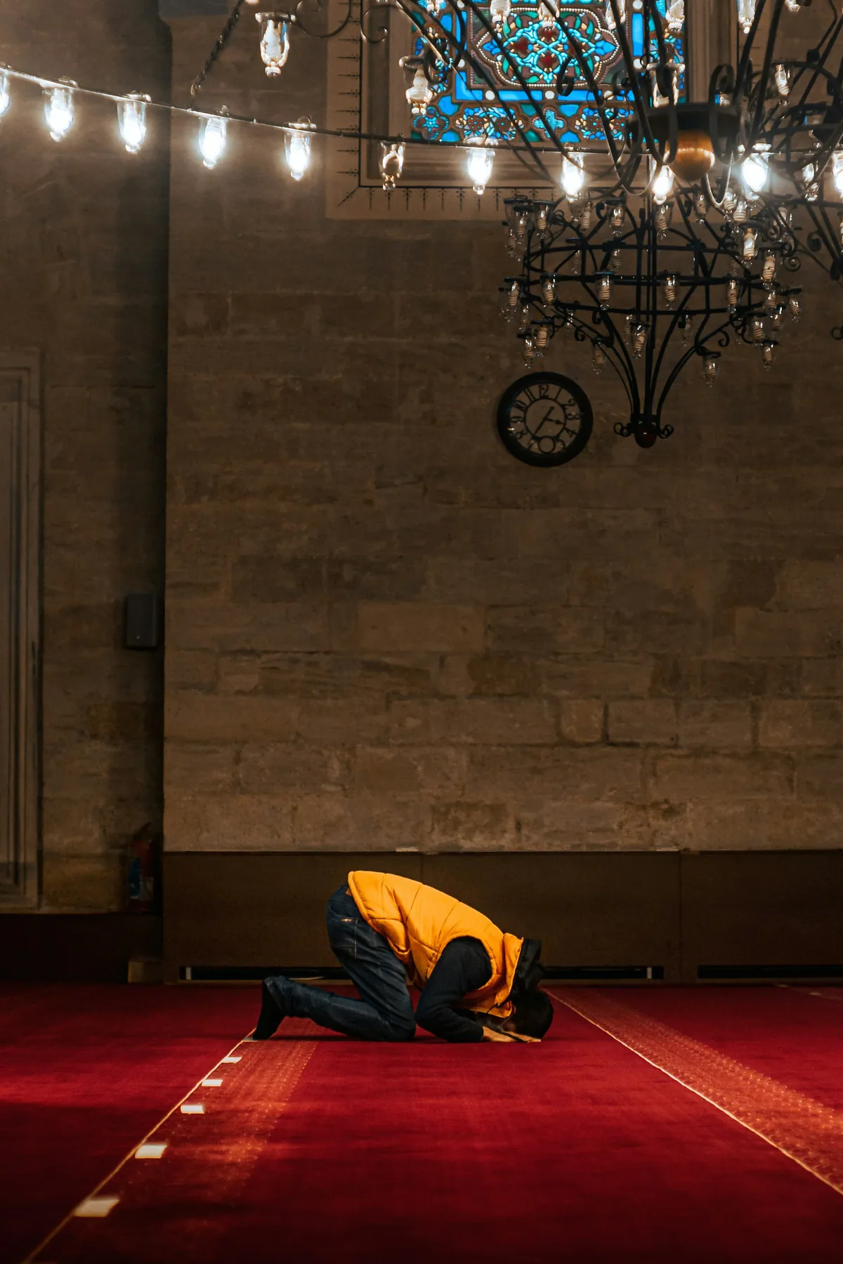 A person in a yellow jacket prostrating in prayer on a red carpet beneath hanging lamps.