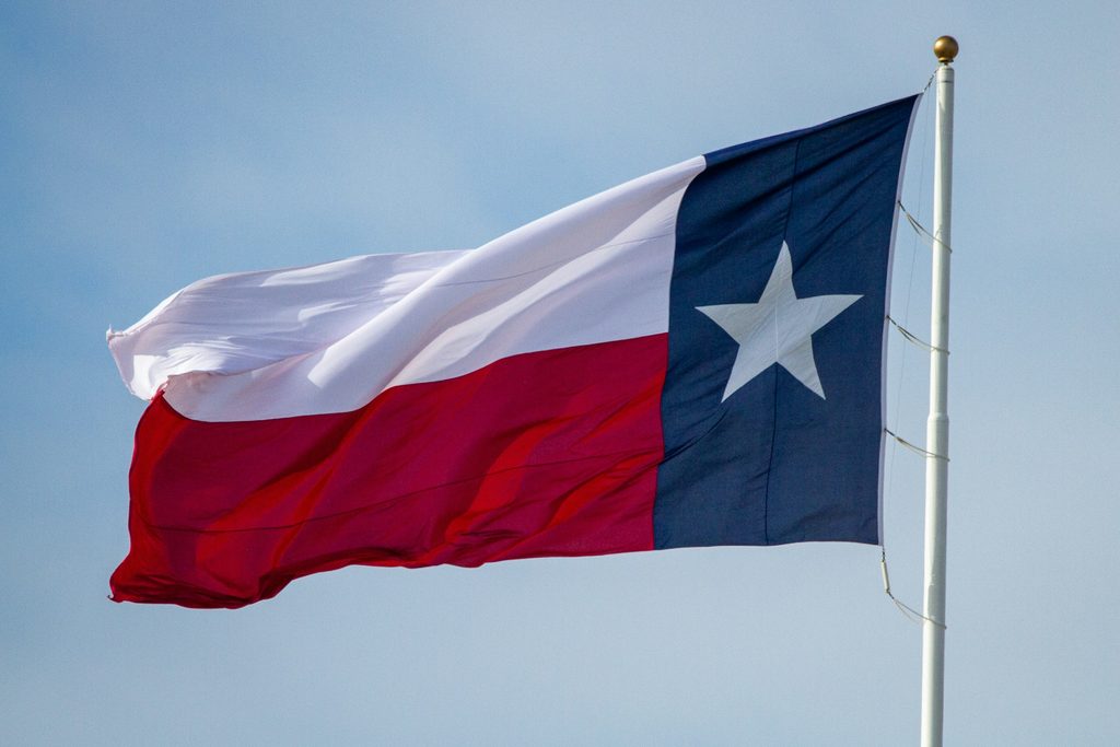 A Texas flag extending to the left from a pole at the right of the frame against a muted blue sky.